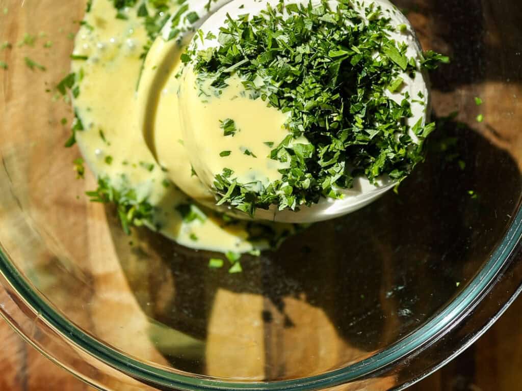 A glass bowl containing a food processor filled with a creamy mixture topped with finely chopped fresh herbs, possibly parsley, placed on a wooden surface. The herbs are vibrant green and the mixture is pale yellow.